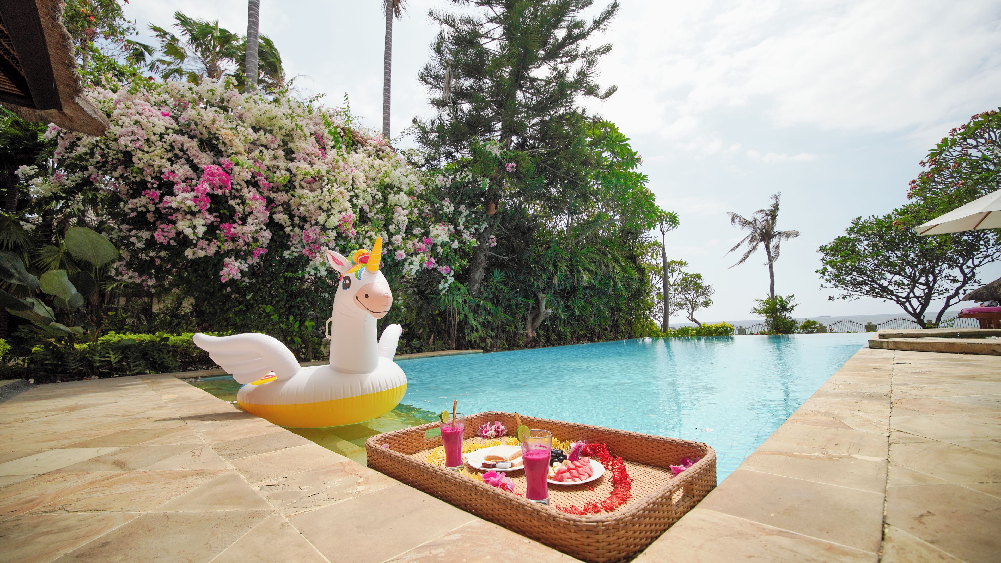 A swimming pool with a unicorn float, a wicker tray with drinks and snacks, and lush greenery.