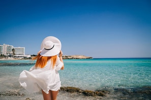 A person standing at the beach with a clear view of the sea and buildings in the background. - Napa Central Apartment (Ayia Napa)