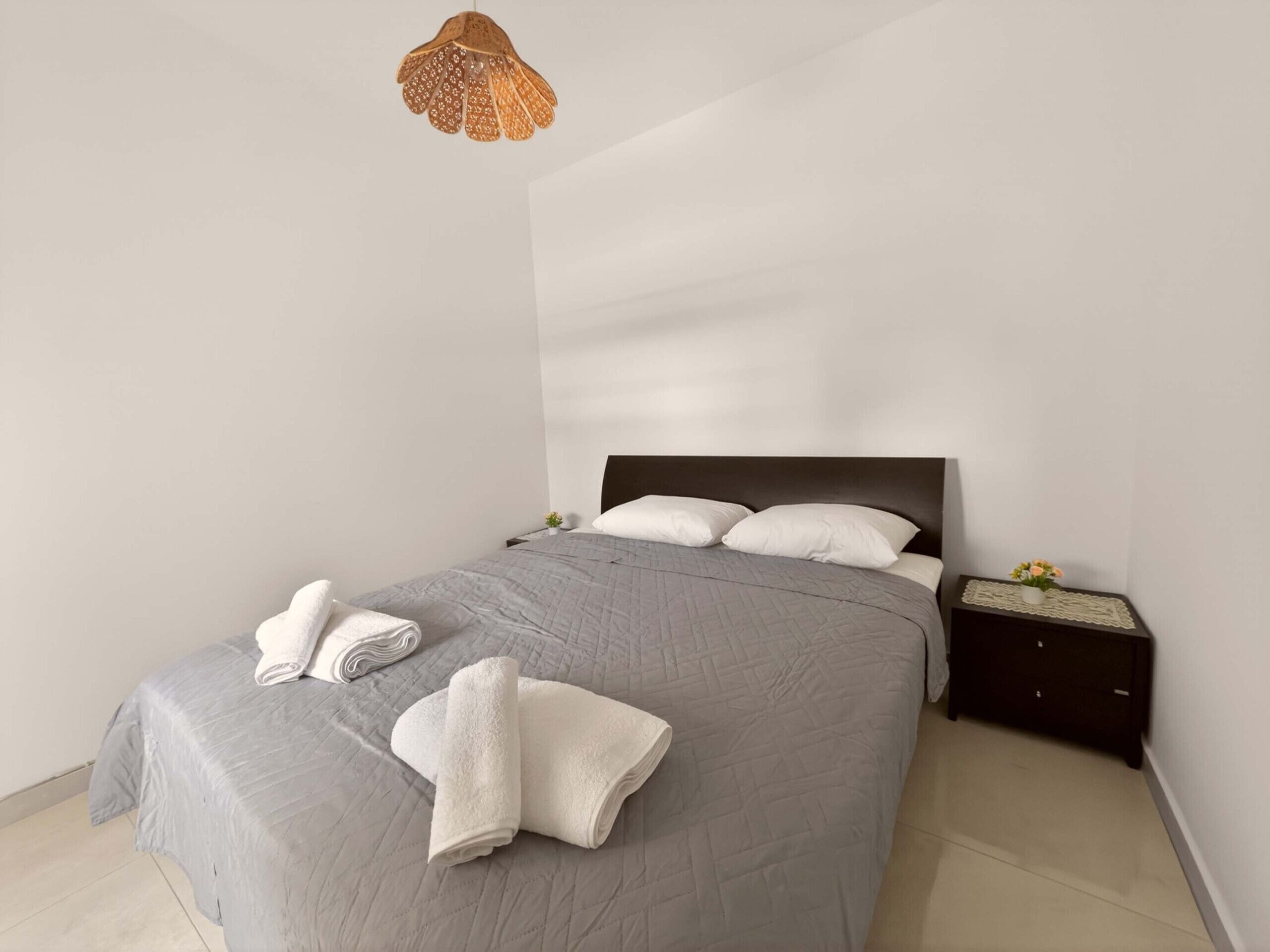A neatly made bed with a grey quilt, two white towels, a wooden nightstand with a small plant, and a hanging light fixture.