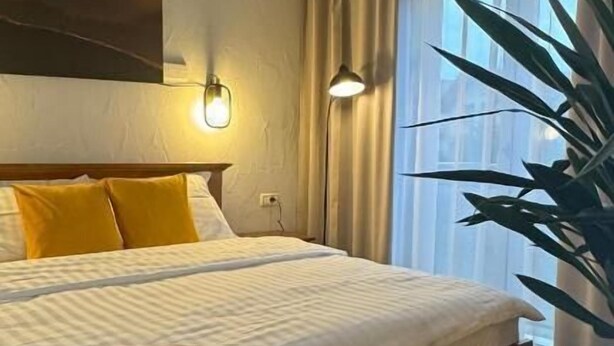 Ein Hotelzimmer mit einem großen Bett, einem Wandbild, einer Topfpflanze und einem Fenster mit Vorhängen.