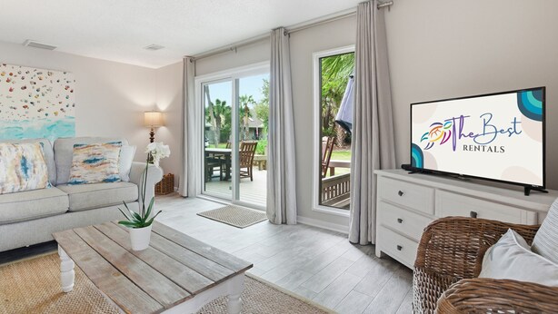 Una sala de estar moderna con un sofá, una mesa de centro con una planta, un televisor sobre un mueble bajo y un gran ventanal corredizo que da a un patio exterior.
