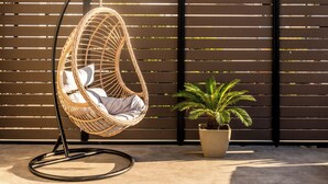 Una silla colgante de mimbre con cojines, una planta de palma en maceta y un muro de listones de madera.