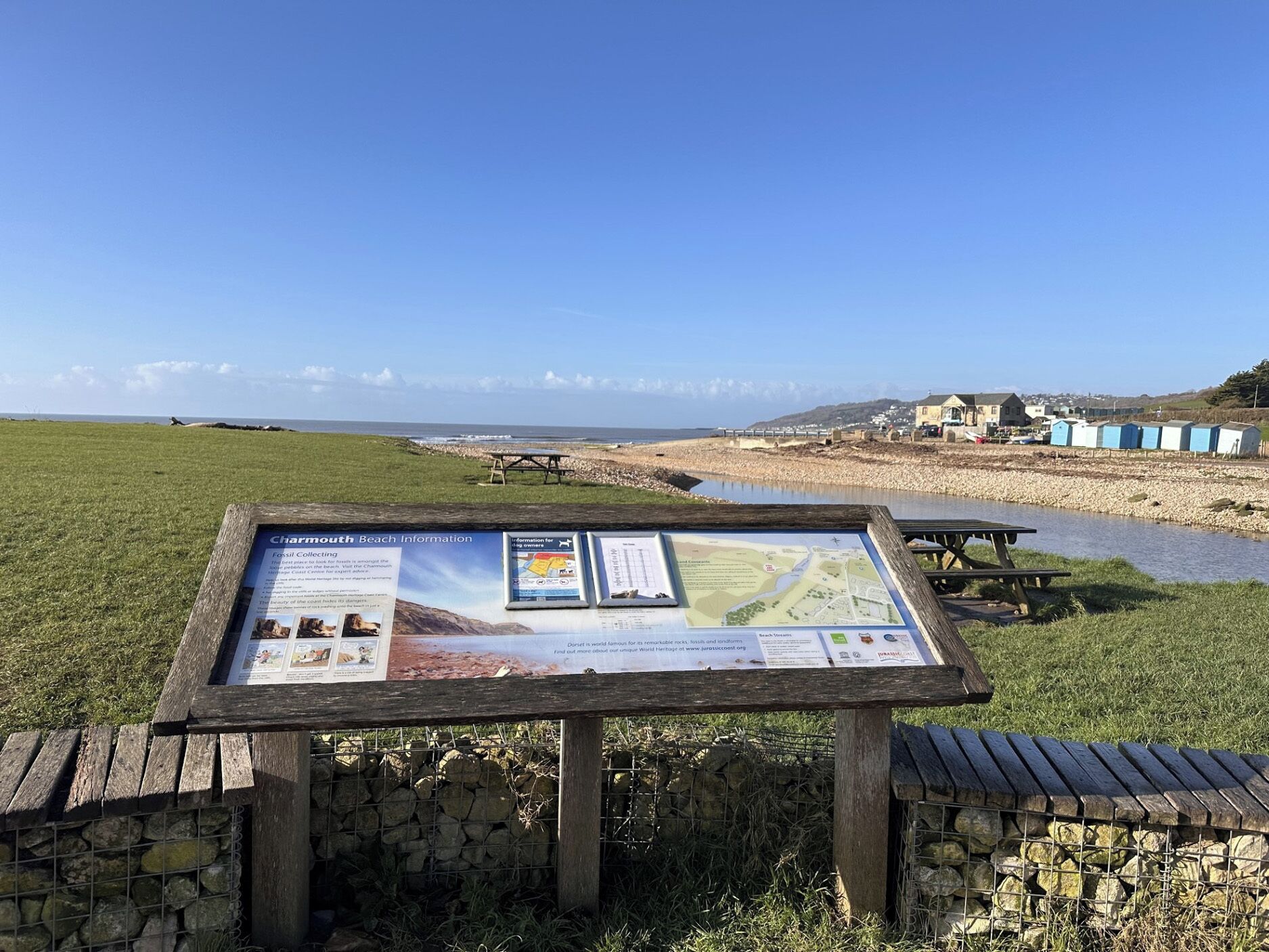 Um painel de madeira com mapas e texto, com o cenário costeiro e céu limpo ao fundo.