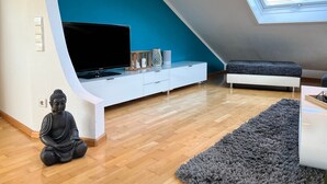 A modern living room with a sloped ceiling, a flat-screen TV, a meditation statue, a sofa, and a large window. - Apartment “Black Forest Brandenkopfblick” with Terrace, Garden and Wi-Fi (Zell)
