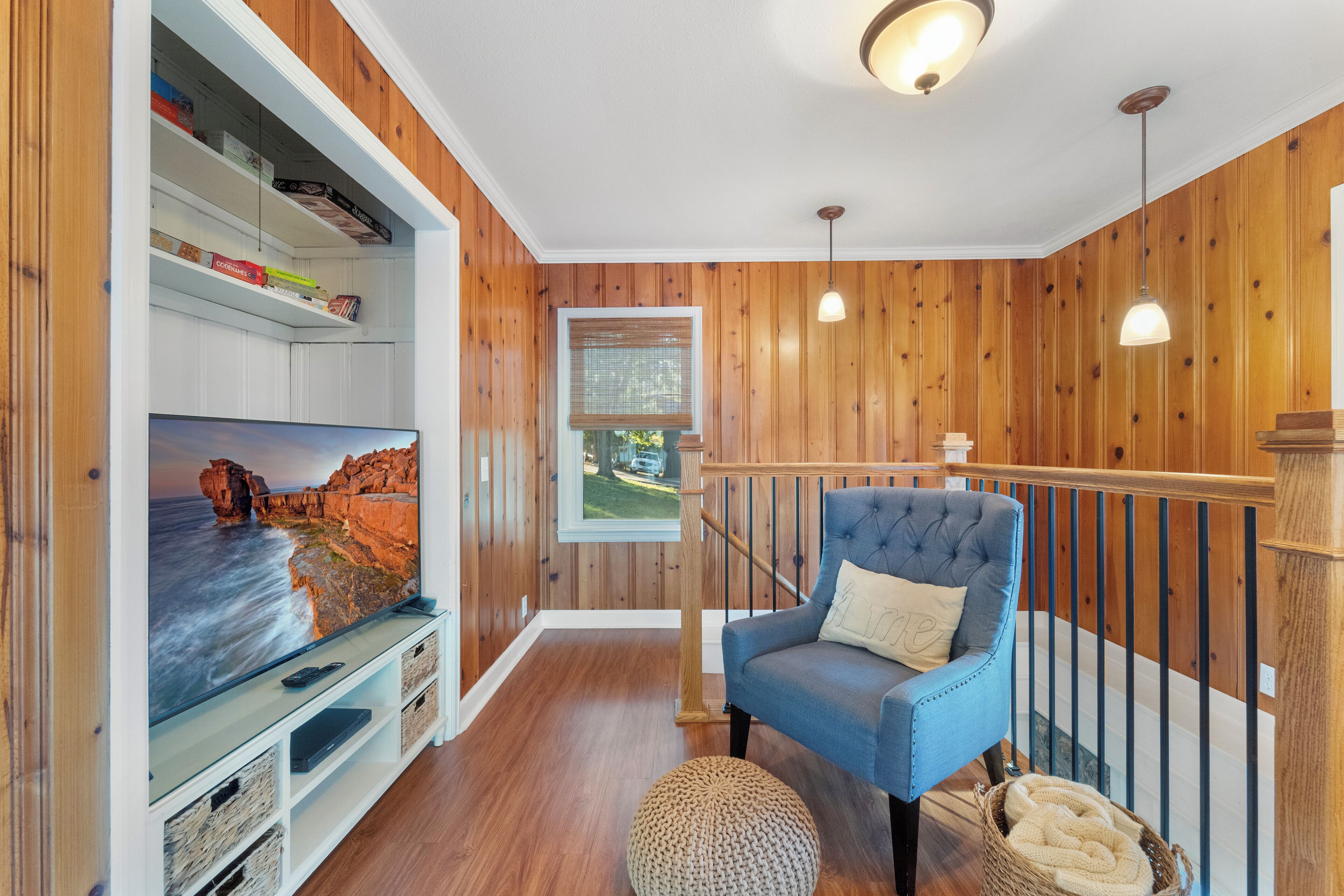 Ein Zimmer mit Holzwänden, einem blauen Sessel, einem Fernseher auf einer weißen Kommode und einem Fenster mit Blick auf Grünflächen.