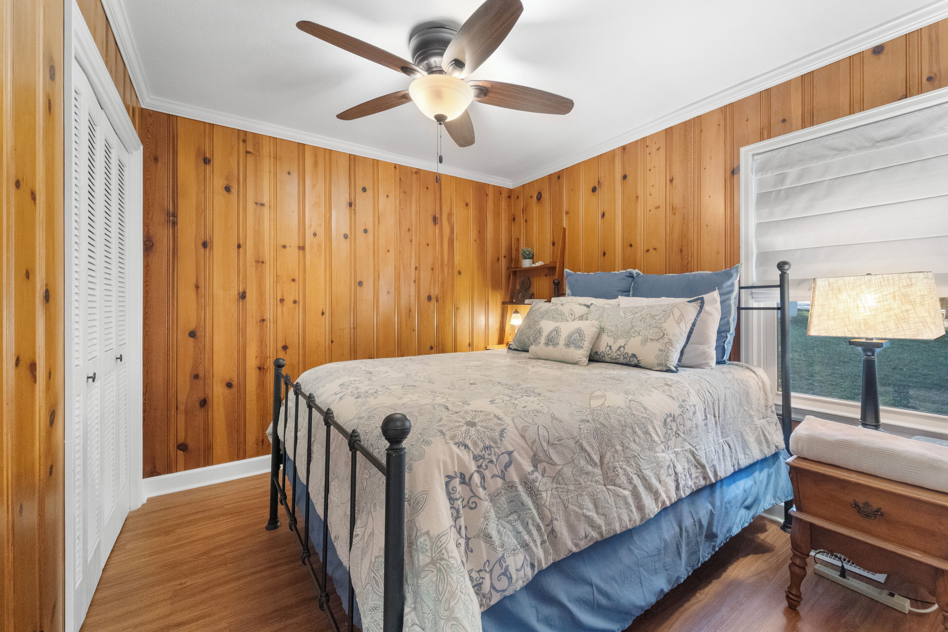 A bedroom with wooden paneling, a bed with patterned bedding, a ceiling fan, a nightstand with a lamp, and a window with a view.