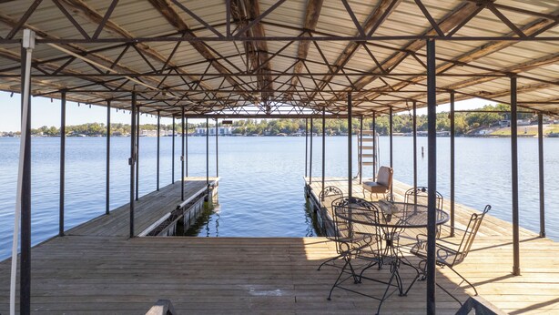 Una terraza techada al aire libre con piso de madera, sillas de metal y una mesa, con vistas a una masa de agua tranquila.