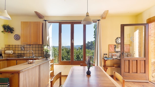 A kitchen with wooden cabinets, a dining table, and a view through the windows. - Holiday Home 'Cal Malla Petit' with Mountain Views, Private Pool & Wi-Fi (Artes)