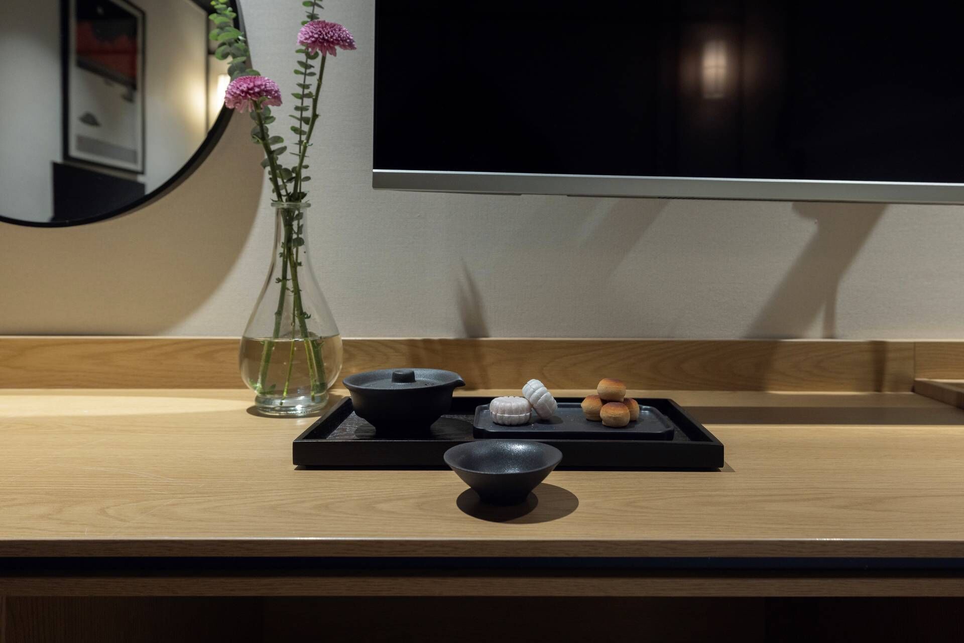 A wooden table with a black tray holding a bowl, a small black container, and some round pastries. A clear glass vase with pink flowers is on the table.