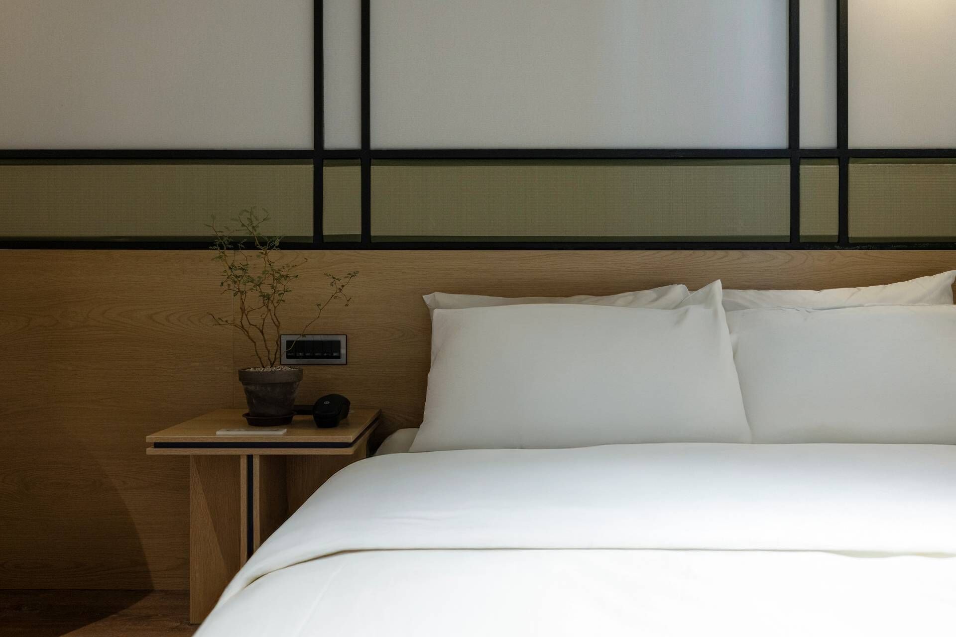 A neatly made bed with white linens in a room with a wooden headboard and a small bedside table with a potted plant.