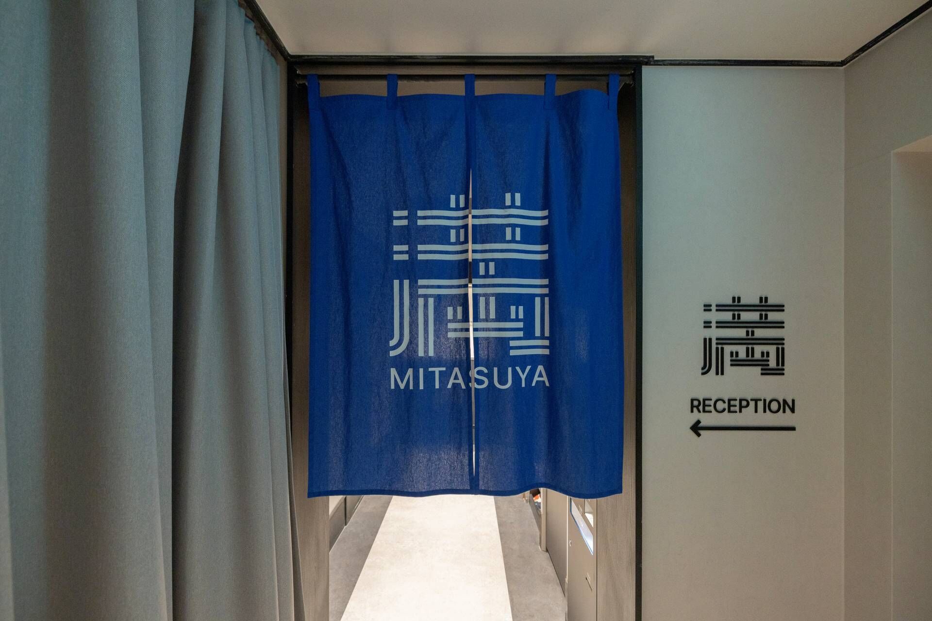 A hotel reception area with a blue curtain displaying the name 'MITASUYA' and a sign indicating the direction to the reception.