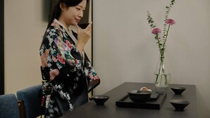 Een persoon in een kimono zittend aan een tafel met een bloevas en traditionele Japanse theeaangelegenheden.