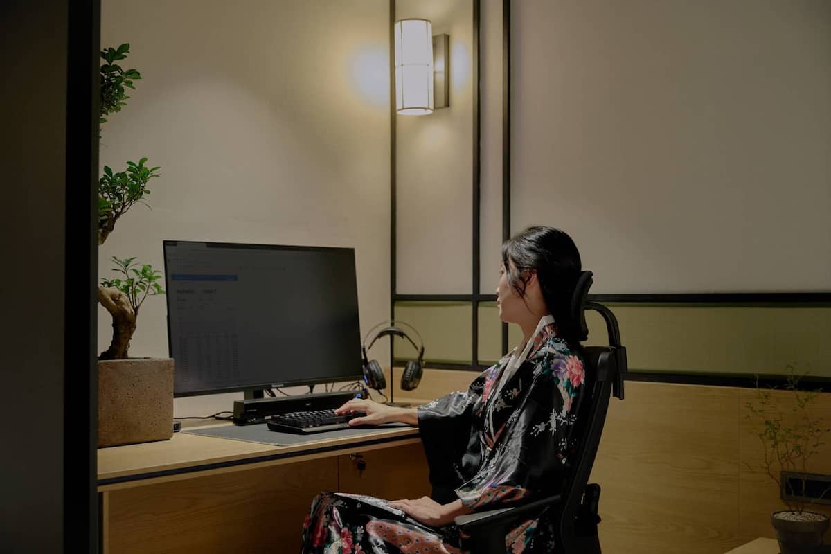 Eine Person sitzt an einem Schreibtisch mit einem Computer, einer Topfpflanze und einer Wandlampe.