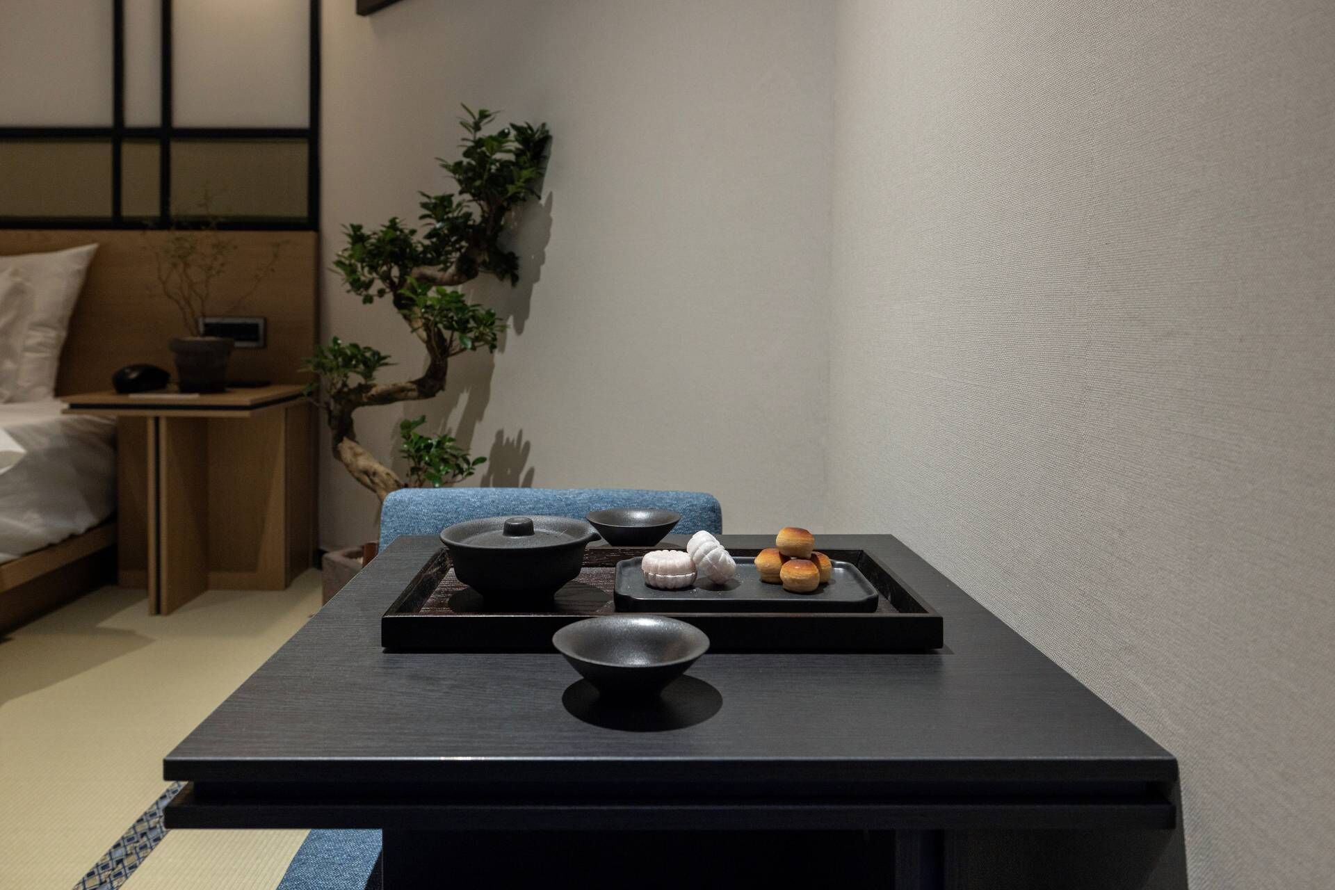 A minimalist Japanese-style room with tatami flooring, a low table with a black tray holding a bowl, a small bowl, and a few pillows.