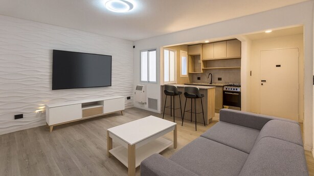 Ein modernes Wohnzimmer mit Flachbildfernseher, einer Couch und einem Couchtisch. Es gibt eine Küchenzeile mit Herd, Spüle und Barhocker.