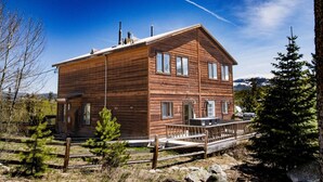 Una baita di legno a due piani con un balcone, immersa tra gli alberi e con montagne sullo sfondo.