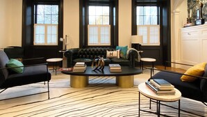 Una sala de estar con un sofá de cuero negro, una mesa de centro redonda y un tapete con estampado de cebra. Hay tres ventanas con persianas, un candelabro y un aparador blanco con objetos decorativos.