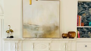 Una habitación con un sillón azul, una mesa redonda con una copa y botella de vino, una chimenea y un cuadro abstracto grande sobre la repisa.