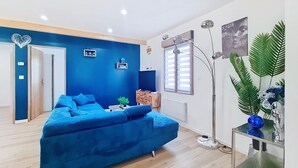 Un salon moderne avec un canapé bleu, une table basse et un lampadaire. Une porte donne accès à une autre pièce et une fenêtre est équipée de stores.