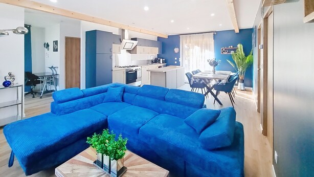 Un salon moderne avec un grand canapé d’angle bleu, un coin repas comprenant une table et des chaises, et une cuisine dotée de meubles blancs et d’un four encastré.