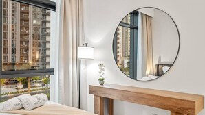 Camera d'albergo con una grande finestra, uno specchio rotondo, un tavolino consolle in legno, un letto con coperta beige e una piccola orchidea in vaso.