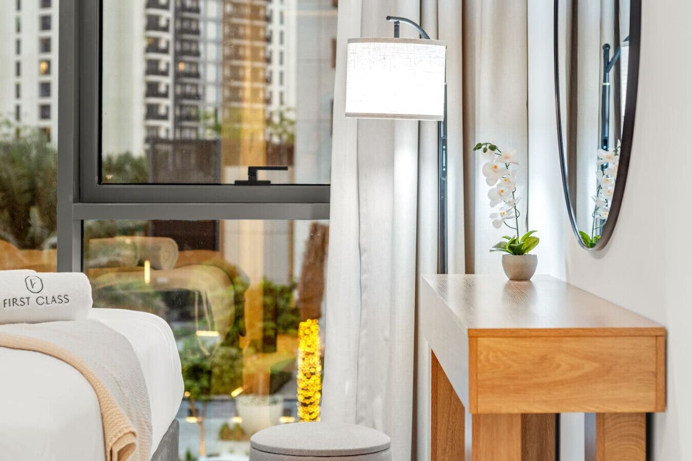 Habitación de hotel moderna con cama, mesita de noche con planta, espejo y ventana con vistas a la ciudad.