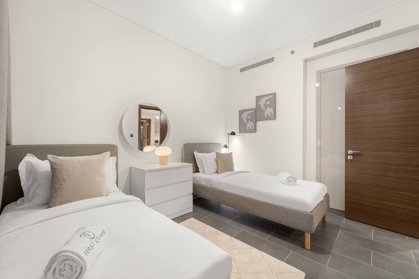 Habitación de hotel con dos camas, un espejo redondo, lámparas de noche, una puerta de madera y una alfombra pequeña.