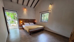 A bedroom with a bed, wooden headboard, two wall-mounted lights, a window with a metal grill, and a view of greenery outside. - JOE BANANA STAYS (Anjuna)