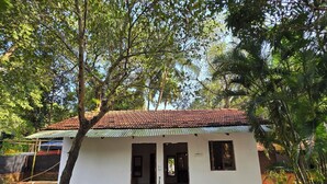 A small white building with a tiled roof, surrounded by trees and a paved area with steps leading to the entrance. - JOE BANANA STAYS (Anjuna)