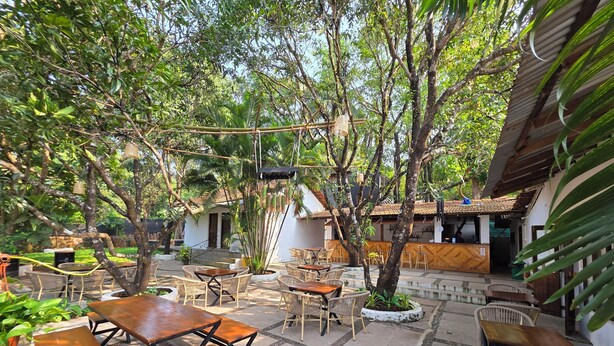 An outdoor patio with wooden tables and wicker chairs, surrounded by lush greenery and trees. - JOE BANANA STAYS (Anjuna)