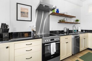 Una cocina moderna con una campana de acero inoxidable, lavavajillas y estufa. Hay repisas de madera sobre la encimera con vajilla y una planta.