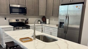 A modern kitchen with a marble countertop, stainless steel appliances, and dark wooden stools. - Coastal Haven-New 3BR/2BA Home, near Beaches+Fenced Yard, Pet-Friendly! (Pensacola)