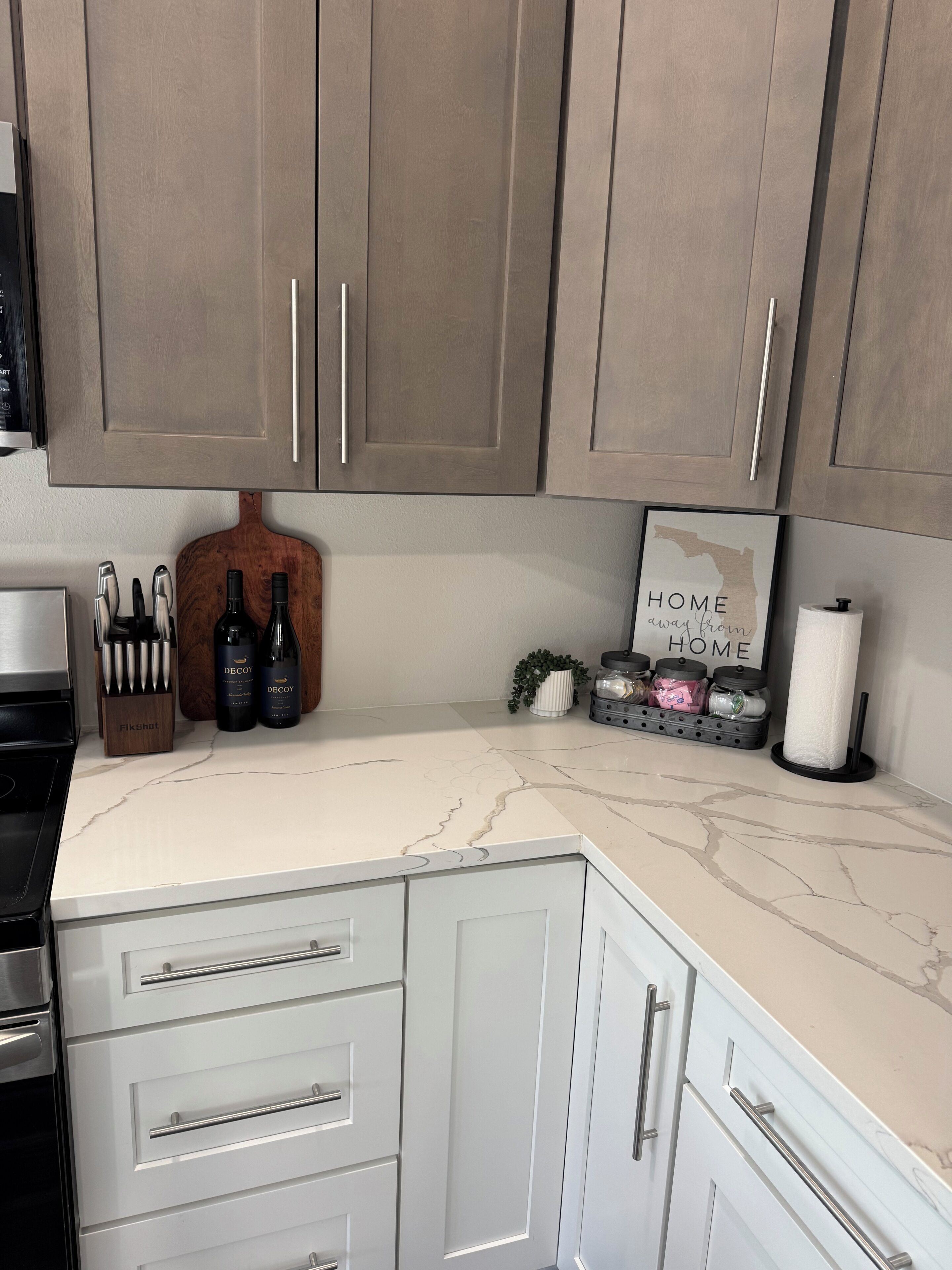 Una cocina con armarios blancos, encimera de mármol y un letrero que dice 'HOGAR lejos del HOGAR'.