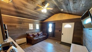 Ein geräumiger Raum mit Holzbalkendecke, Deckenventilator, einer Couch, einem Fernseher und einer Tür mit Fenster.