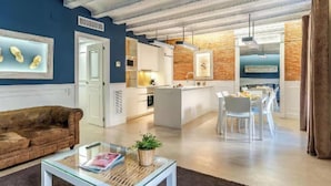 Een moderne woonkamer met een bank, een glazen salontafel en een keukenhoek met witte kasten en een bakstenen muur.