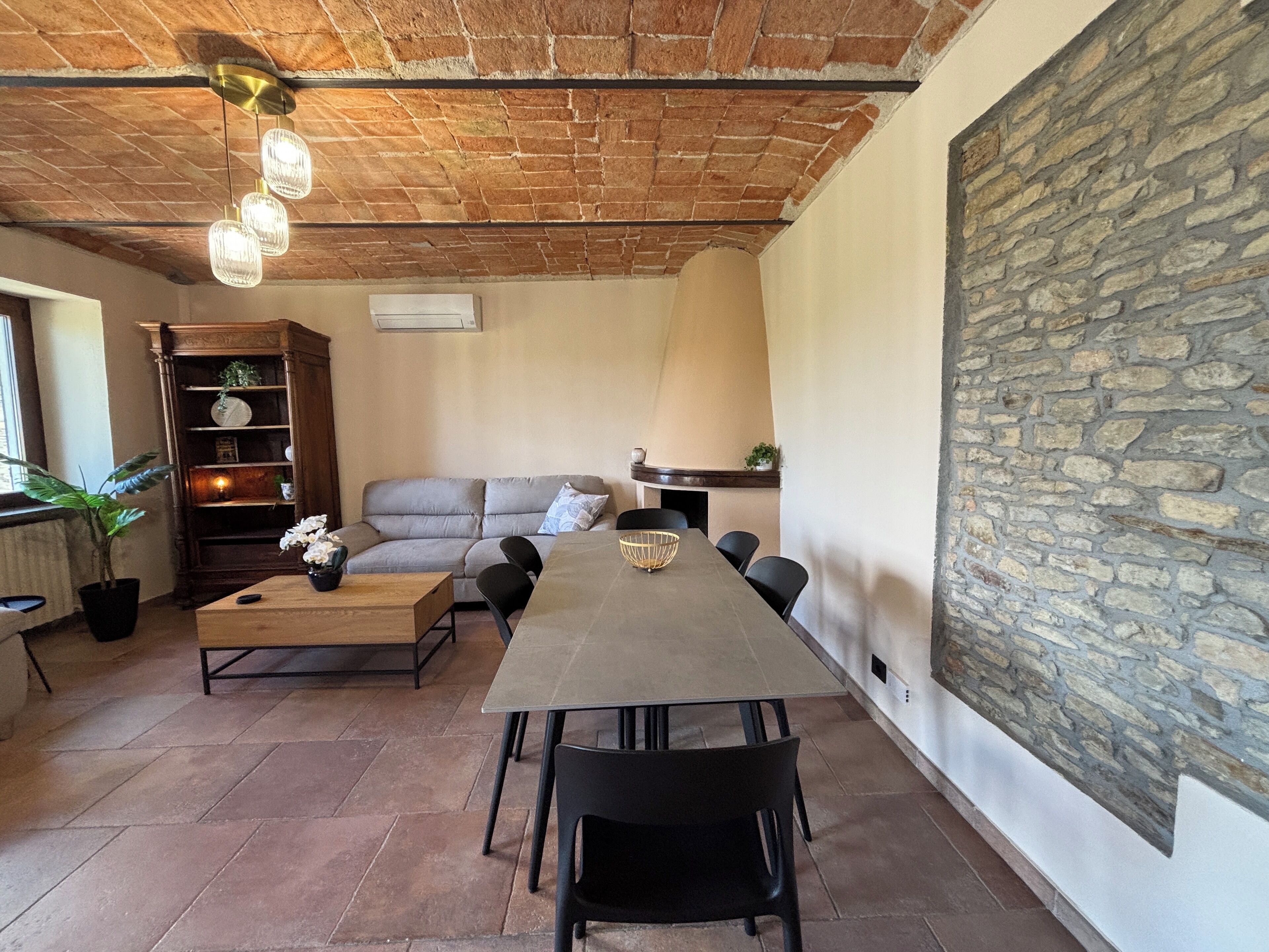 A spacious living area with a long dining table, black chairs, a grey sofa, and a stone wall.