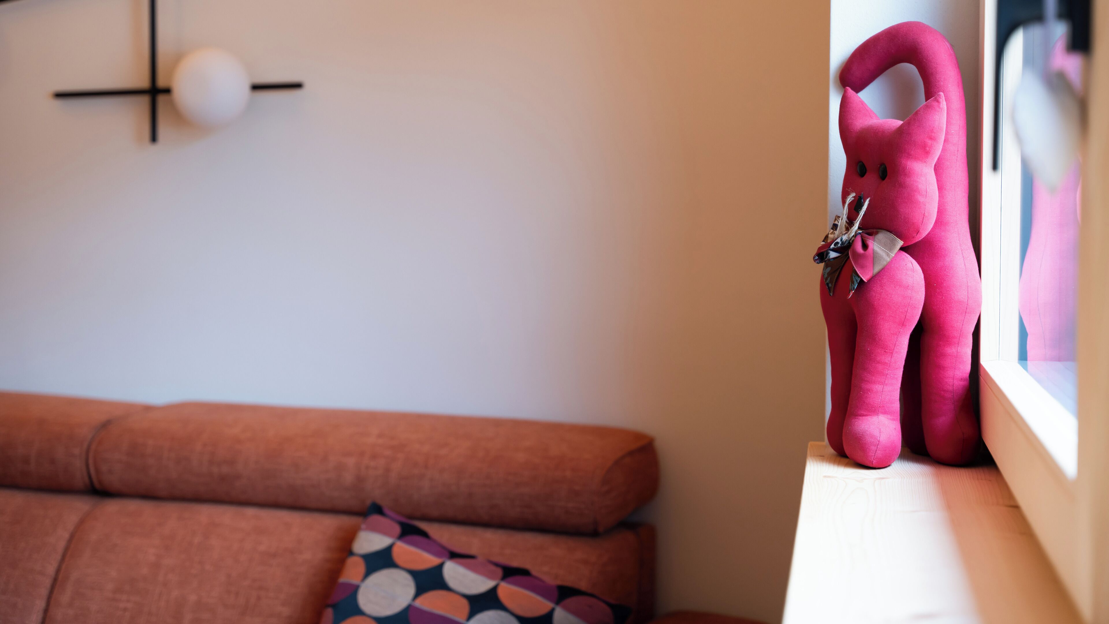 Un salon chaleureux avec un canapé marron, un coussin à motifs et un ours en peluche rose posé sur un rebord de fenêtre.