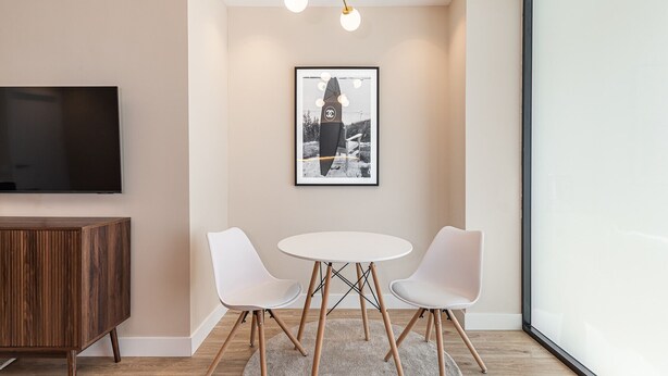 Ein modernes Zimmer mit Fernseher, einem gerahmten Bild, zwei weißen Stühlen, einem runden Tisch und einem Holzkasten.