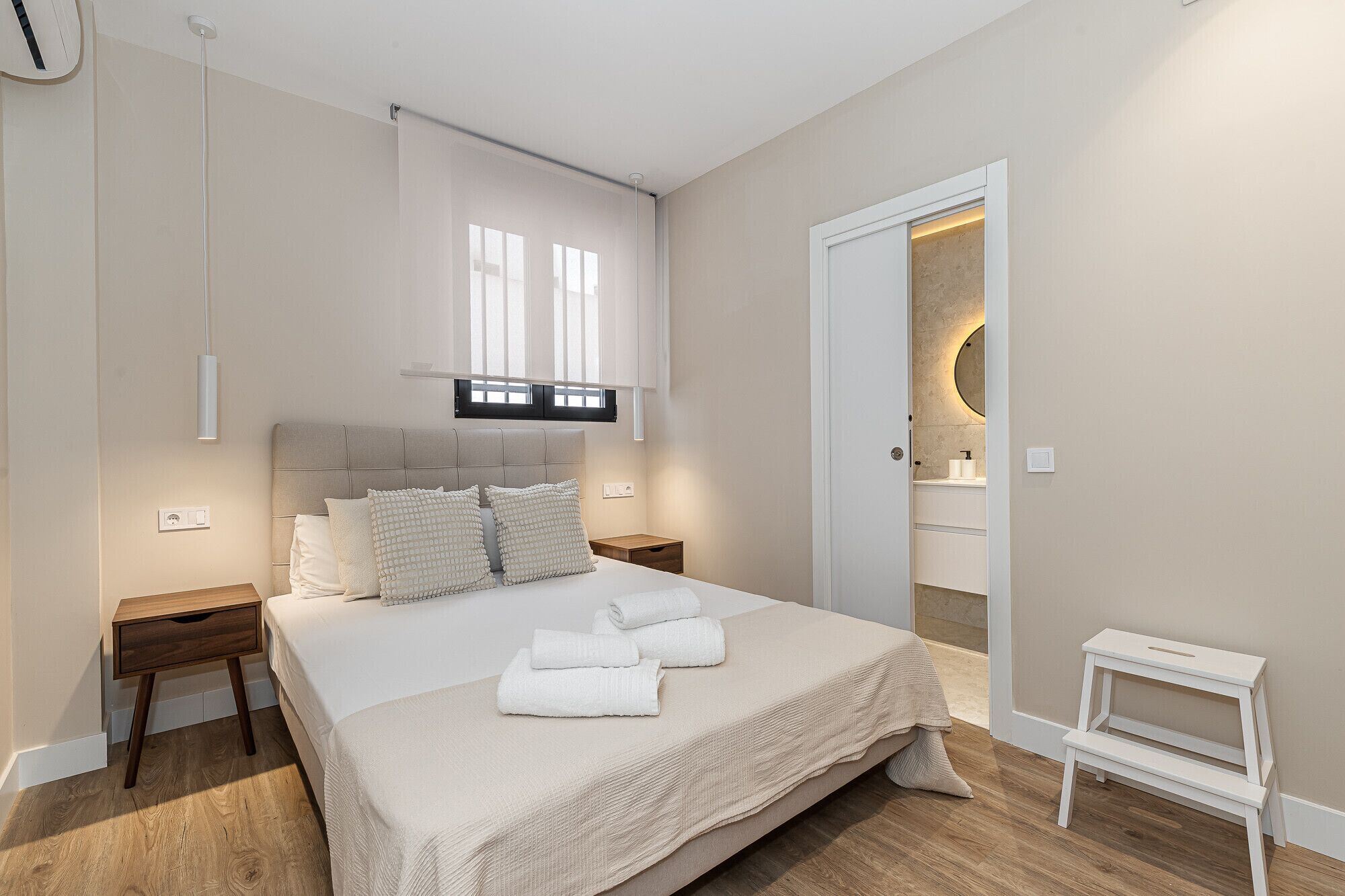 A modern bedroom with a large bed, bedside tables, a bedside lamp, a door leading to a bathroom, and a small white stool.