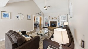 Un salon spacieux avec un haut plafond, une cheminée et un coin repas visible à travers une porte ouverte.