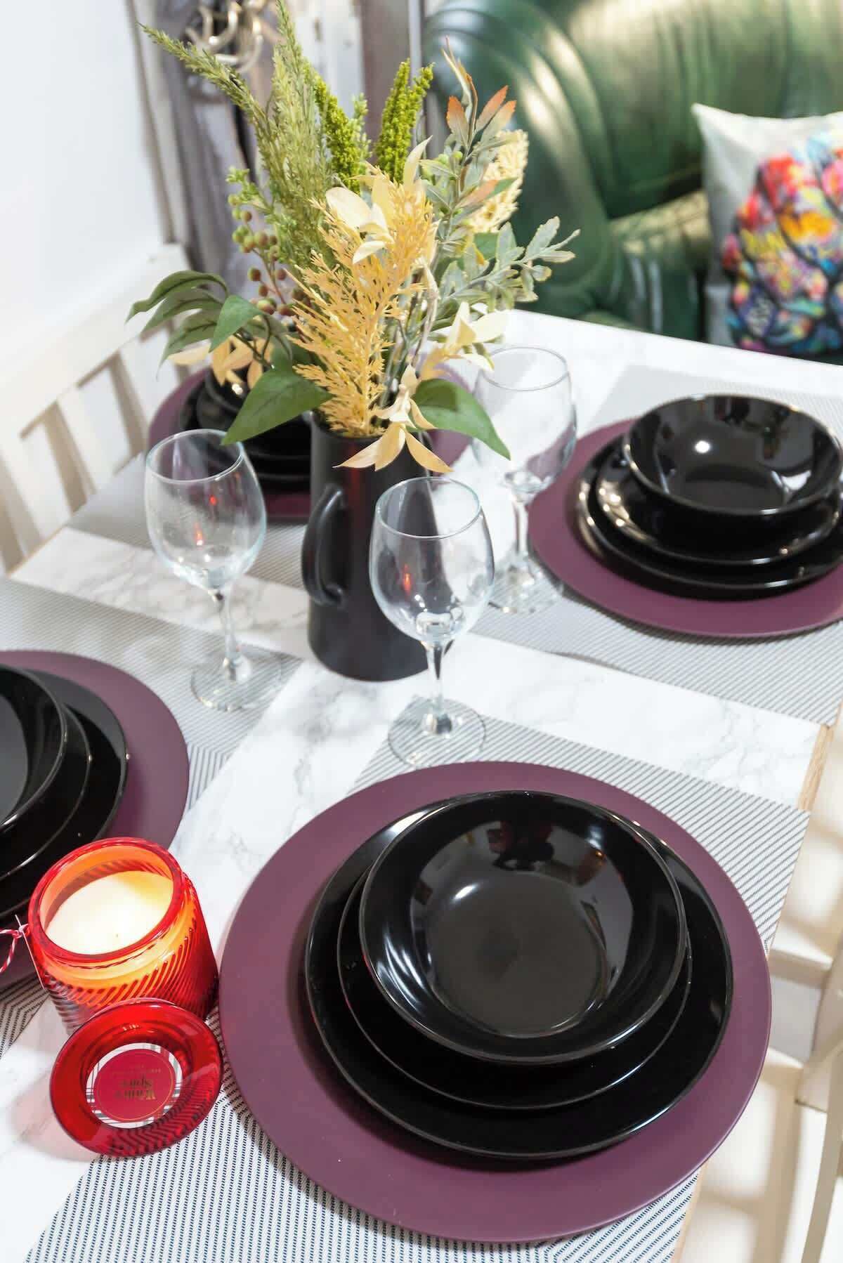 Ein gedeckter Tisch mit schwarzen Tellern, lilafarbenen Servietten und klaren Gläsern, ergänzt durch einen Blumenstrauß und eine brennende Kerze.