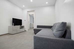 Un salon moderne avec un canapé gris, un téléviseur à écran plat posé sur un meuble en bois, et un couloir menant à une autre pièce.