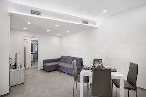A modern, well-lit room with a grey sectional sofa, a dining table with four chairs, and a small white cabinet.
