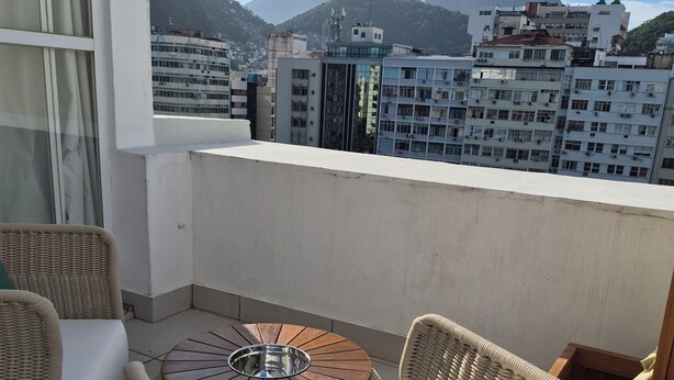 Un balcón con muebles de mimbre, una mesa redonda y vistas a un paisaje urbano con edificios y una montaña.