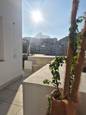 A balcony with a potted plant, a clear view of buildings in the background, and a bright sun. - Penthouse in Copacabana, one block from the beach, with views of the ocean and Christ the Redeemer. (Rio de Janeiro)