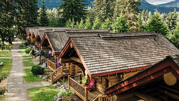 A row of log cabins with wooden decks and railings, surrounded by trees and a mountain backdrop. - Riverside RV Resort (Whistler)