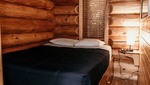 A log cabin room with a bed, wooden walls, a window with blinds, and a bedside table with a lamp. - Riverside RV Resort (Whistler)