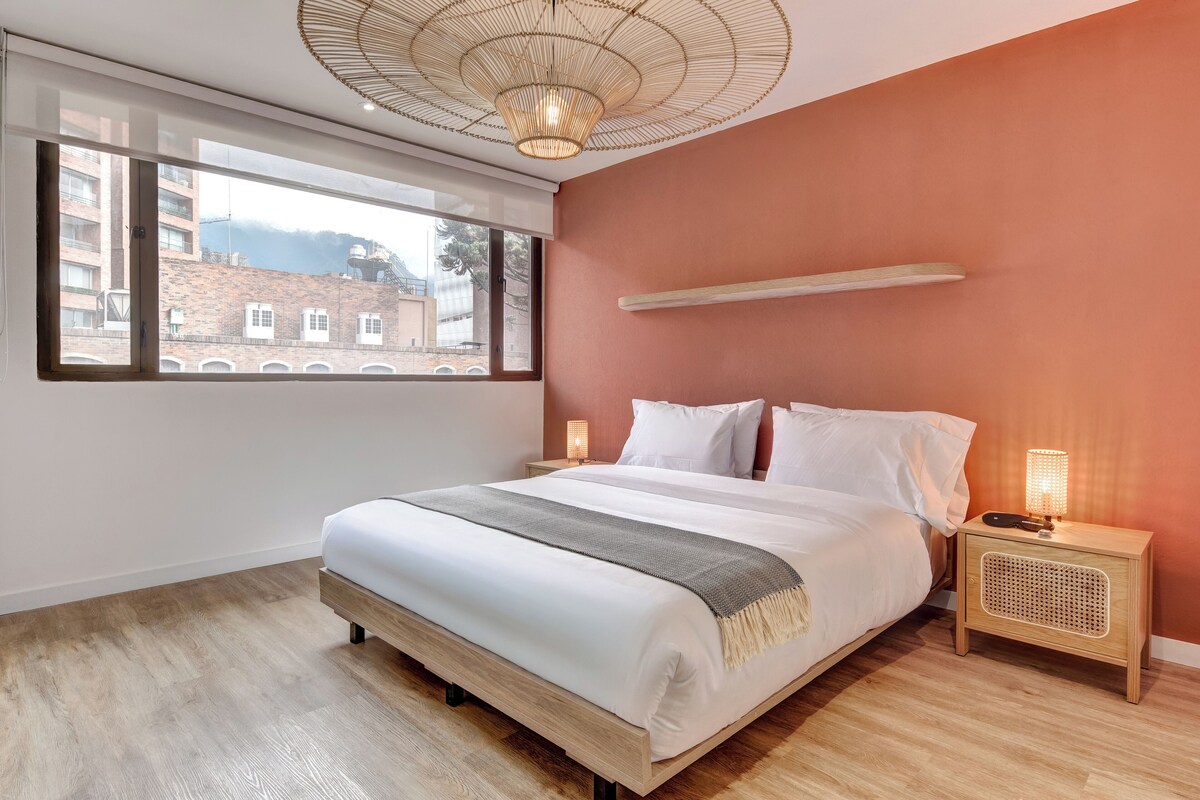 Ein Schlafzimmer mit einem großen Bett, einem Nachttisch mit Lampe, einem Fenster mit Blick auf Gebäude und einem Deckenventilator.