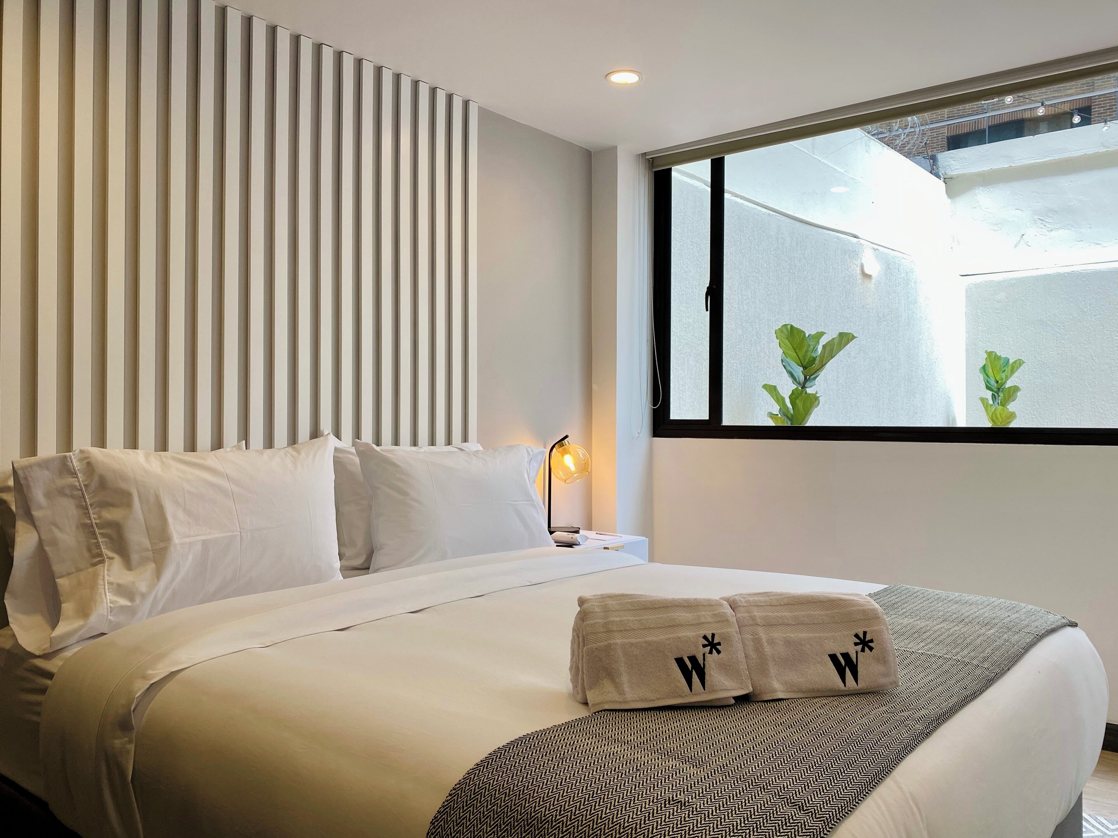 Una cama bien hecha con sábanas blancas, una lámpara de noche y una ventana con vistas a la vegetación.