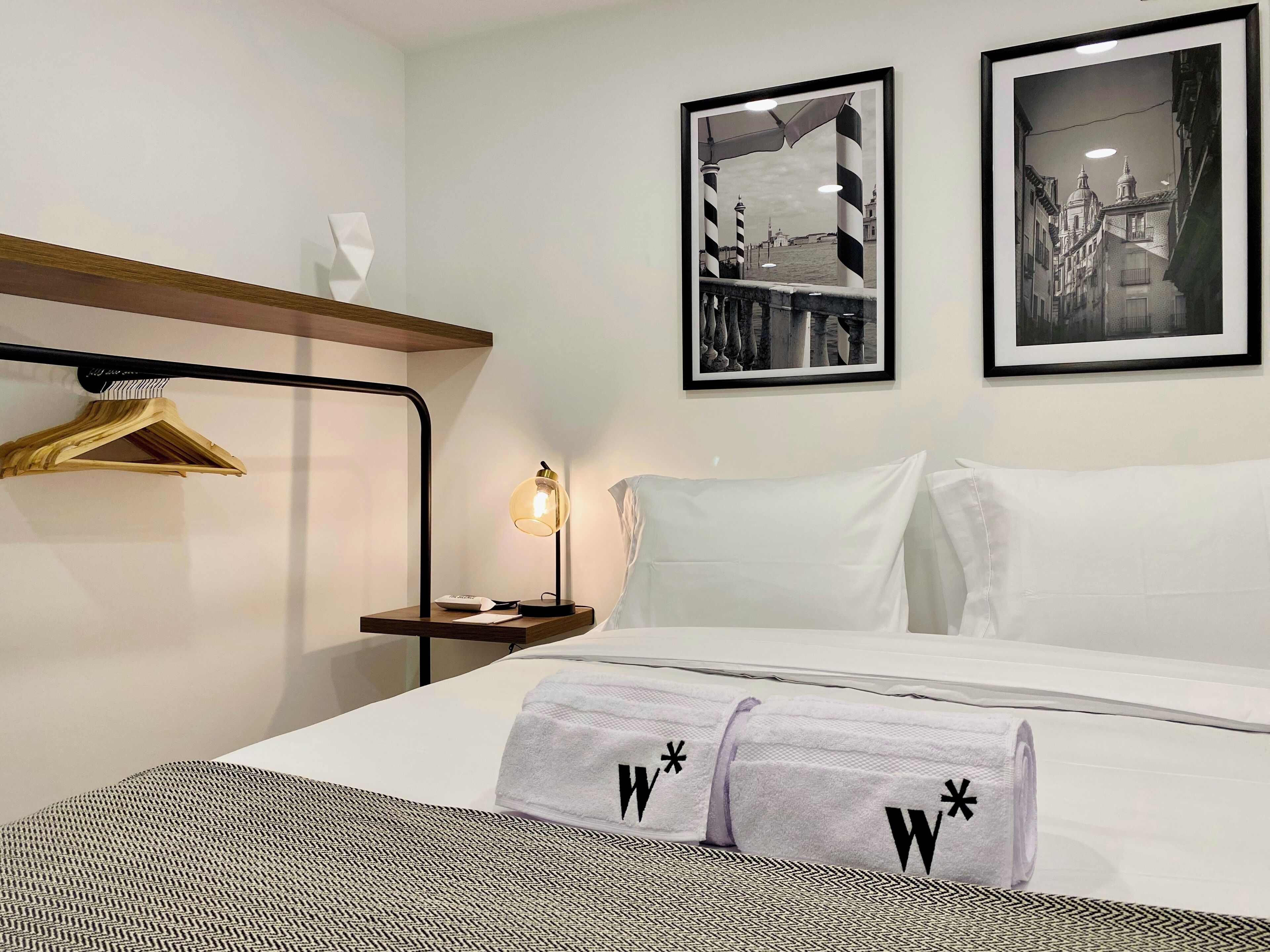Une chambre d’hôtel comprenant un lit, une table de chevet, une lampe, un tableau encadré, un porte-manteau mural et une serviette ornée d’un logo.